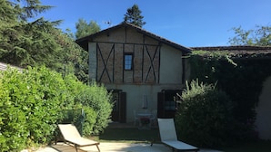 Uma casa de dois andares com piscina, cercada por vegetação e um alpendre com espreguiçadeiras.