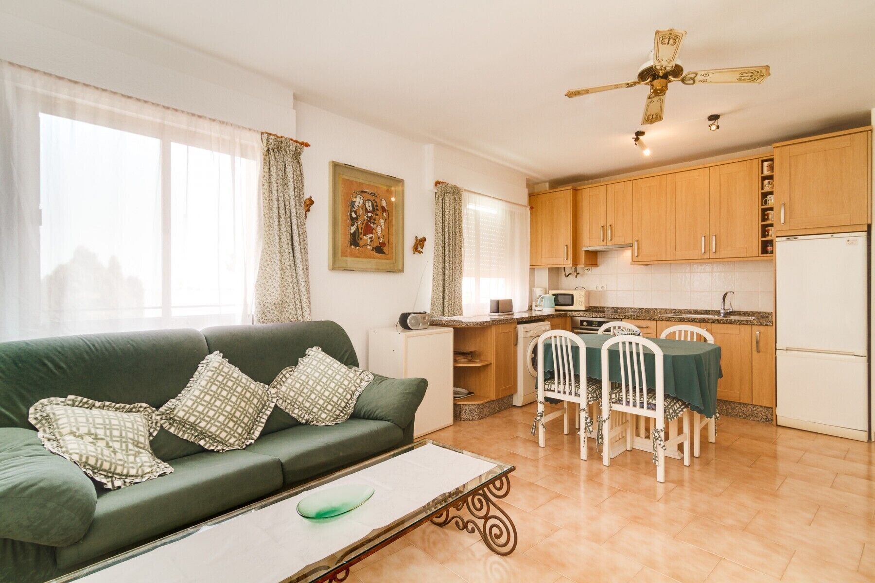 A compact living space with a green sofa, a dining table with chairs, a kitchen area with wooden cabinets, a ceiling fan, and a window with curtains.
