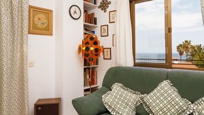 A green sofa with patterned pillows, a bookshelf with books and decorative items, a wall clock, and a window with a view of the sea. - Carabeillo 13 Apartments by Casasol (Nerja)