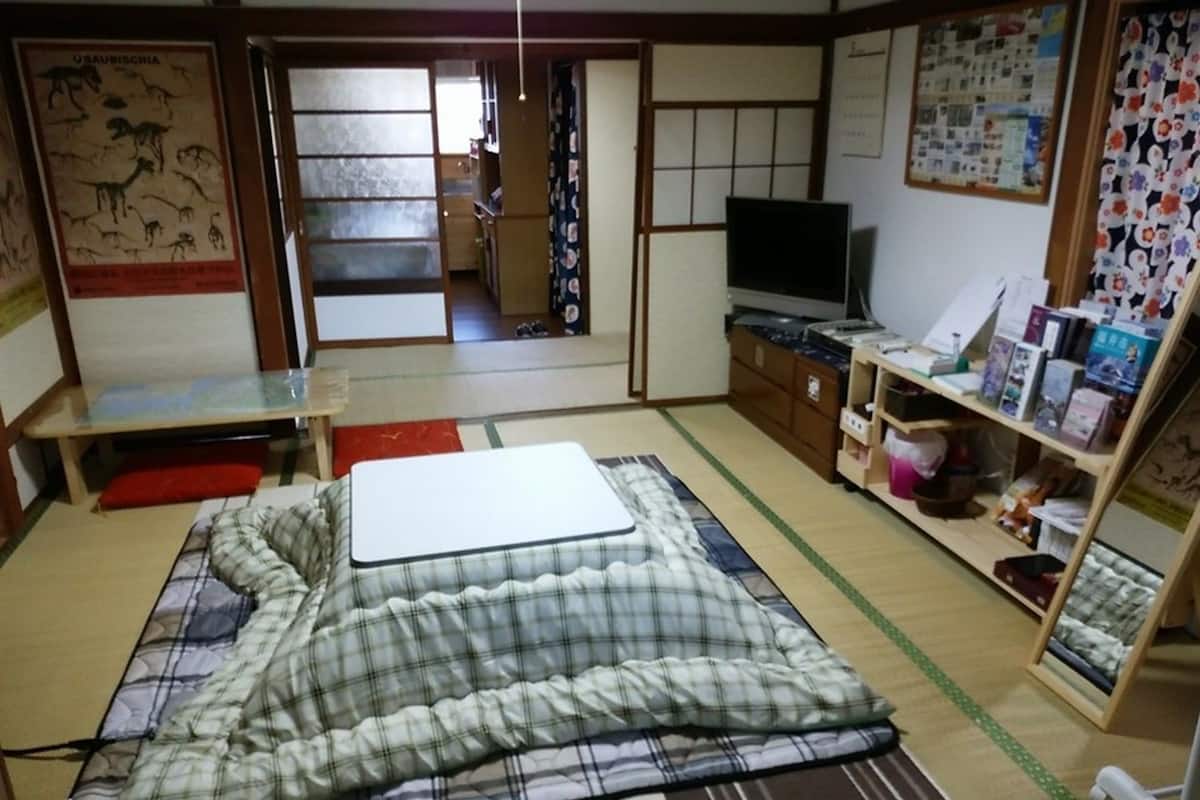 Una habitación japonesa tradicional con suelo de tatami, un futón, una mesa baja, un televisor y una estantería con diversos objetos.