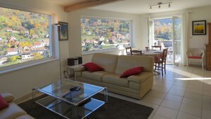 Ein großzügiger Wohnraum mit einem großen Fenster, das einen Blick auf ein Hügeldorf freigibt. Es gibt einen Glastisch, ein beiges Sofa mit roten Kissen sowie eine Essecke mit Tisch und Stühlen.