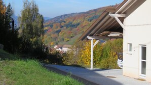 Ein modernes Gebäude mit geneigtem Dach, einer überdachten Außenterrasse und Blick auf ein Tal mit herbstlich gefärbten Bäumen.