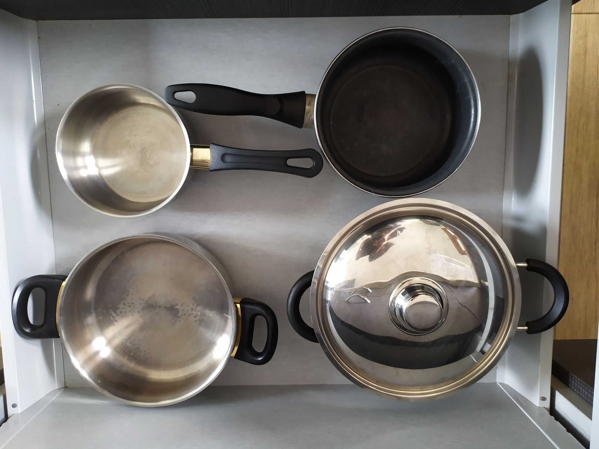 A kitchen with four stainless steel pots and pans, two with black handles and two with gold handles, arranged on a stainless steel countertop.