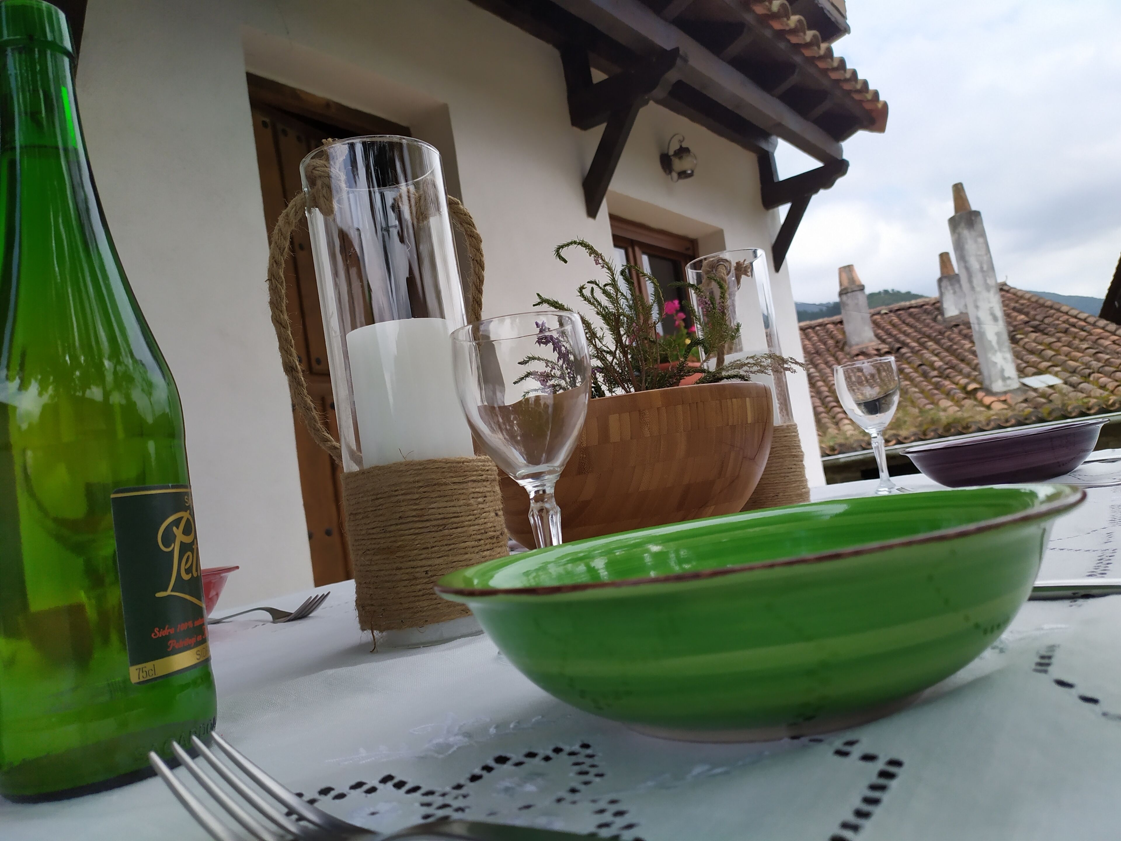 Ein Tisch mit einer grünen Flasche, einem Glas und einer Schüssel. Eine Pflanze steht in einer Holzschüssel, daneben eine weiße Kerze. Der Tisch ist draußen aufgestellt, mit Blick auf ein Haus und ein Ziegeldach.
