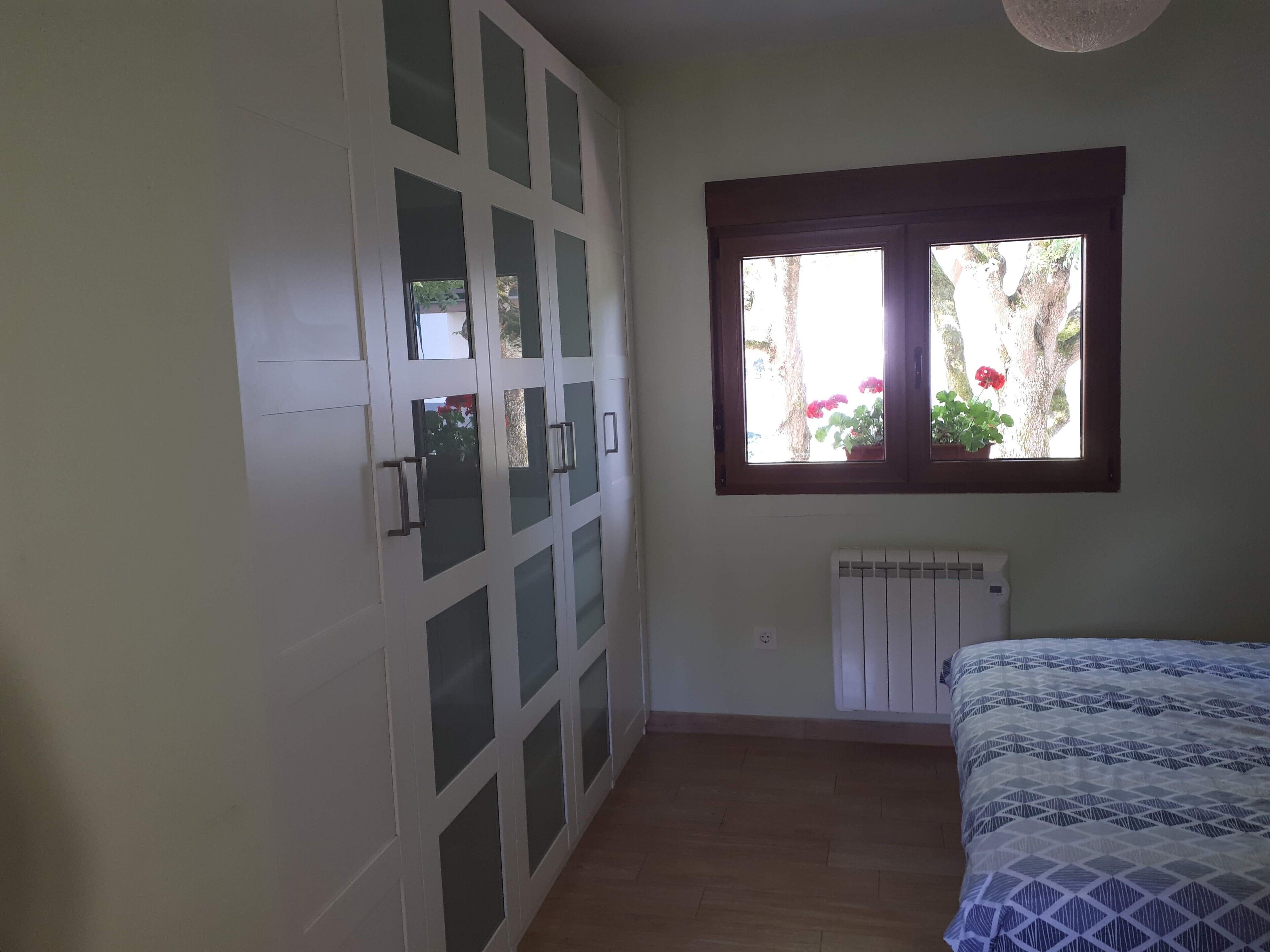A bedroom with a bed, a wardrobe, a window with plants, and a radiator.