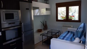 A compact kitchen with a refrigerator, microwave, and oven. There is a small table with a vase and a bowl. A window with a view of greenery is visible. - Rural house with garden and terrace between two natural parks. (Igorre)