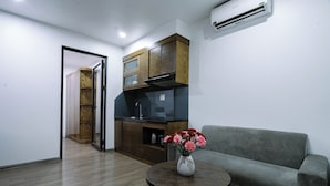 Una sala moderna con un sofá gris, una mesa de centro redonda de madera con un jarrón de flores, una zona de cocina con armarios de madera y un horno empotrado, y una puerta que da a otra habitación.