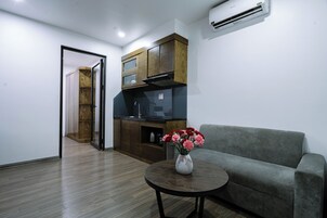 Una sala moderna con un sofá gris, una mesa de centro redonda de madera con un jarrón de flores, una zona de cocina con armarios de madera y un horno empotrado, y una puerta que da a otra habitación.