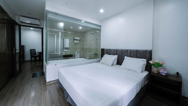 Una habitación de hotel moderna con una cama grande, cabina de ducha de vidrio, piso de madera oscura y un pequeño rincón de descanso.