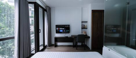 A modern hotel room with a large bed, a desk with a computer, a television, and a glass-enclosed shower.