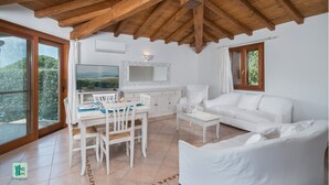 Une pièce spacieuse avec un plafond en bois, un canapé blanc, une table à manger avec des chaises, un téléviseur et une grande fenêtre donnant sur un paysage verdoyant.
