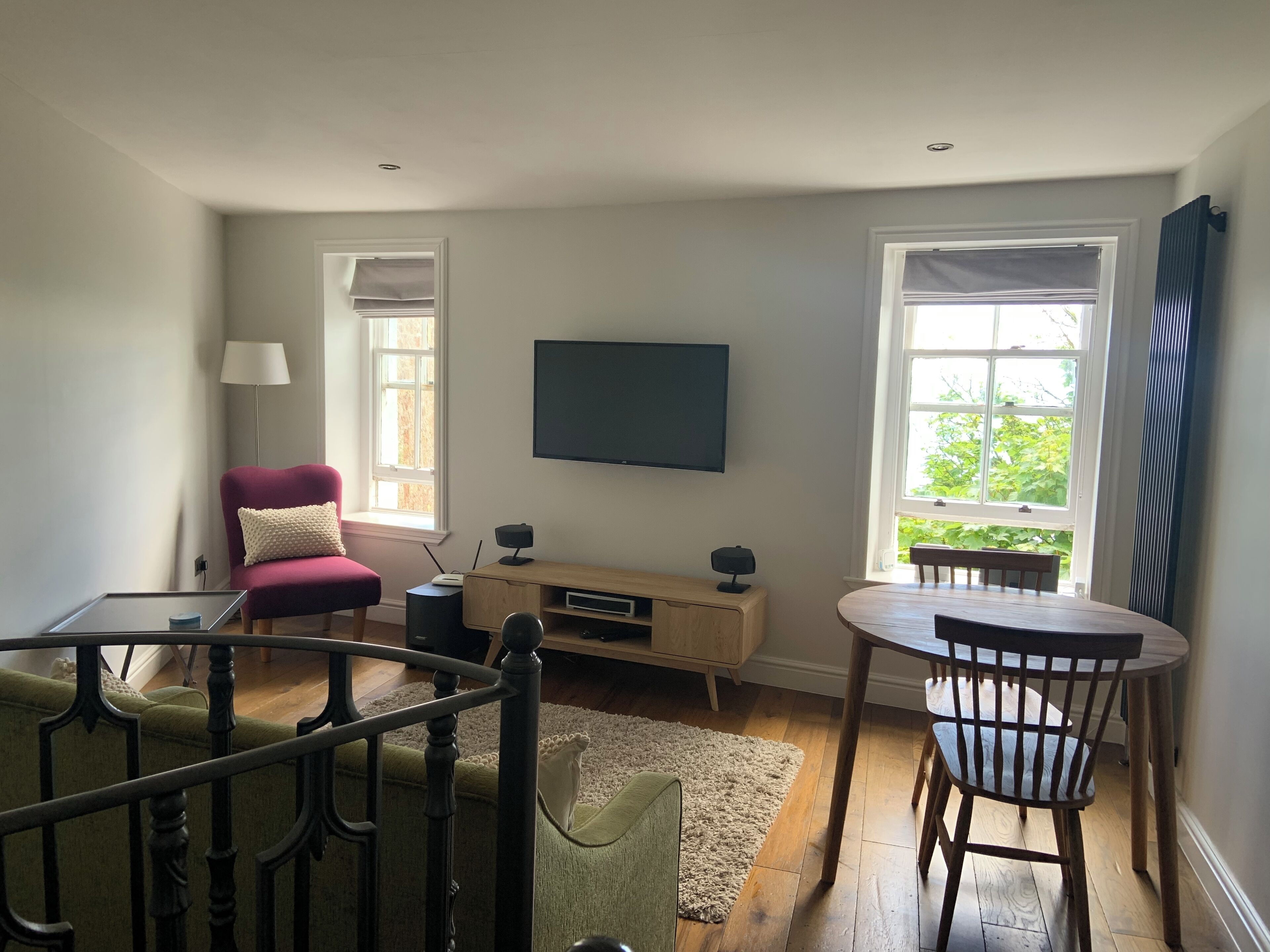 A modern living room with a flat-screen TV, a red armchair, a wooden dining table, and a view of greenery through the windows.