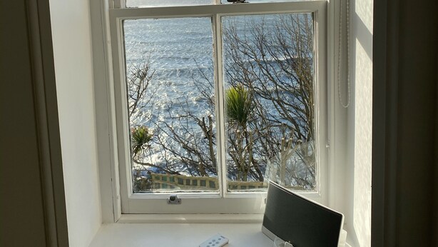 Ein Fenster mit einem Laptop auf einer weißen Fensterbank, mit Blick auf ein Gewässer und kahle Bäume.