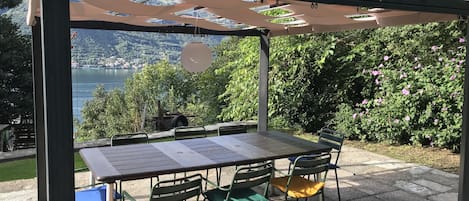 Relax in a serene outdoor seating area under a shaded pergola, surrounded by a tranquil body of water and majestic mountains.