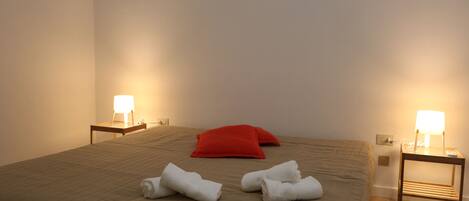 Una cama bien tendida con una almohada roja, dos lámparas de noche y toallas sobre la cama.