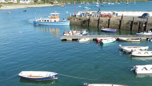 Um porto com vários barcos atracados no cais, uma praia de areia ao fundo e um muro de pedra separando a água da terra.