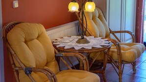 Une pièce avec des fauteuils en osier recouverts d’un tissu jaune, une table ronde avec une nappe en dentelle et deux lampes fixées au mur.