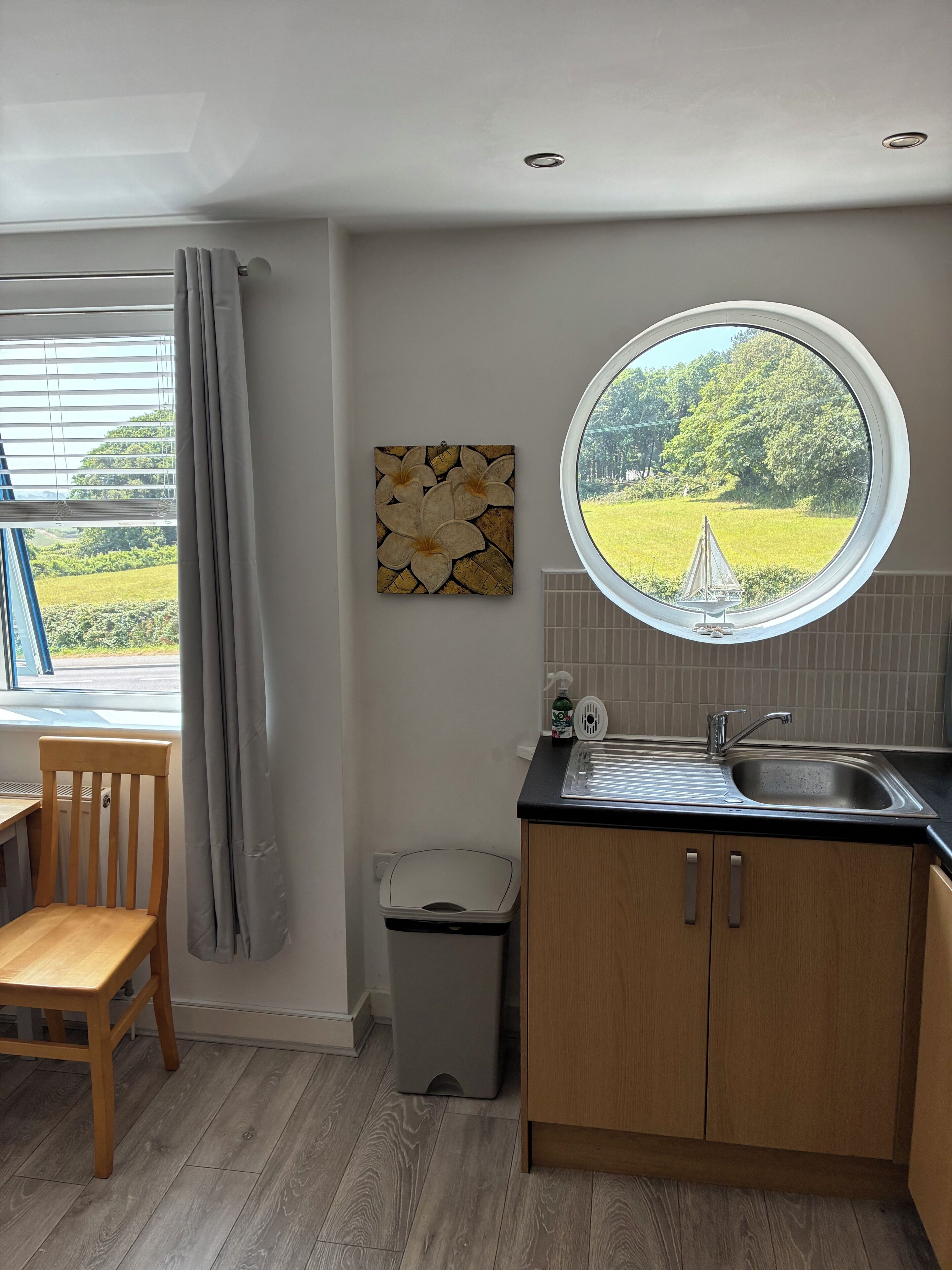 A kitchen with a window, a sink, a chair, and a trash bin.
