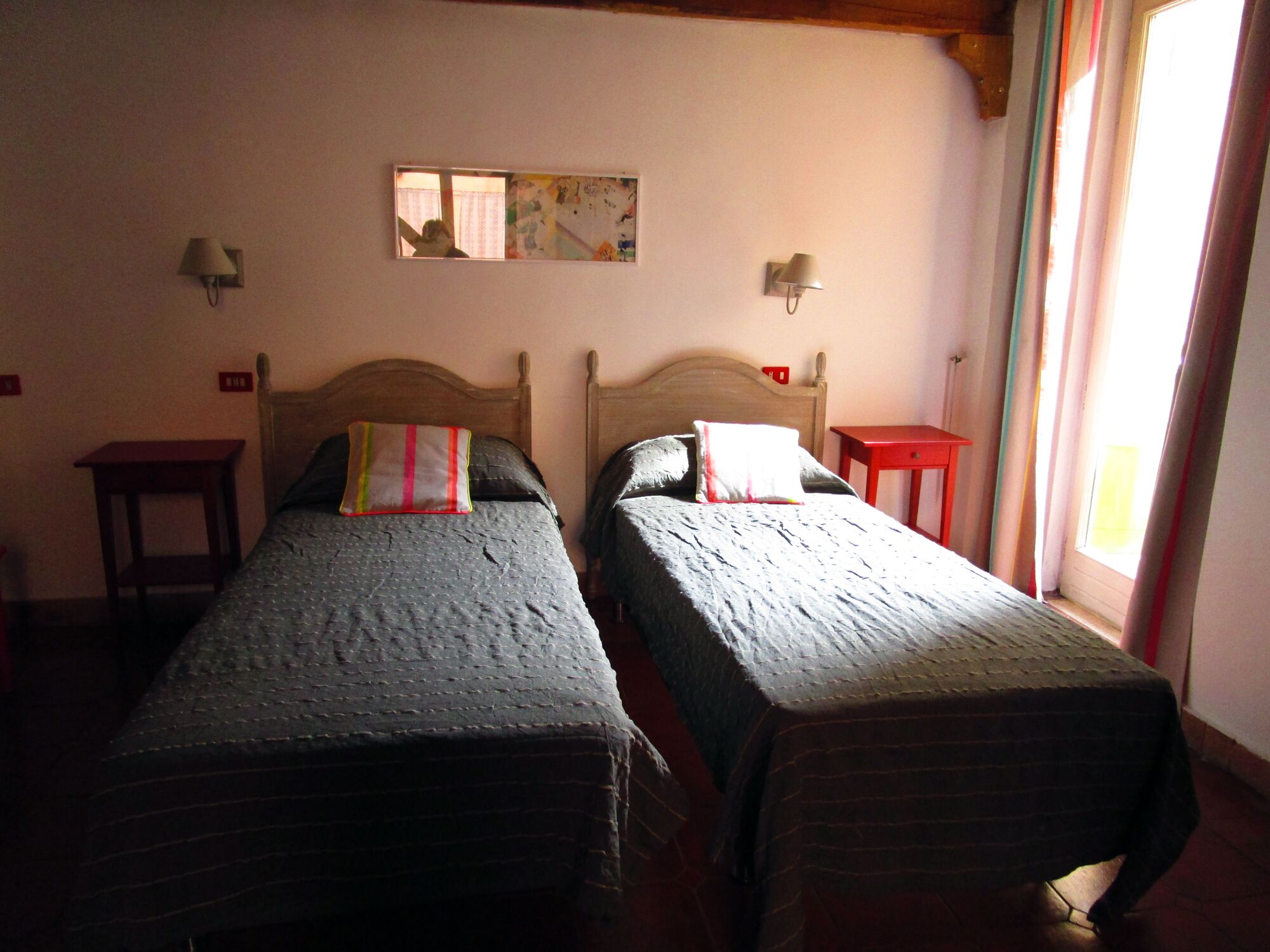 A room with two single beds, each with a pillow and a blanket. There are bedside tables, a wall-mounted lamp, and a window with curtains.