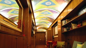A narrow hallway with wooden walls, a tiled floor, and a colorful ceiling. There are bookshelves, a small table, and a couch with colorful cushions.