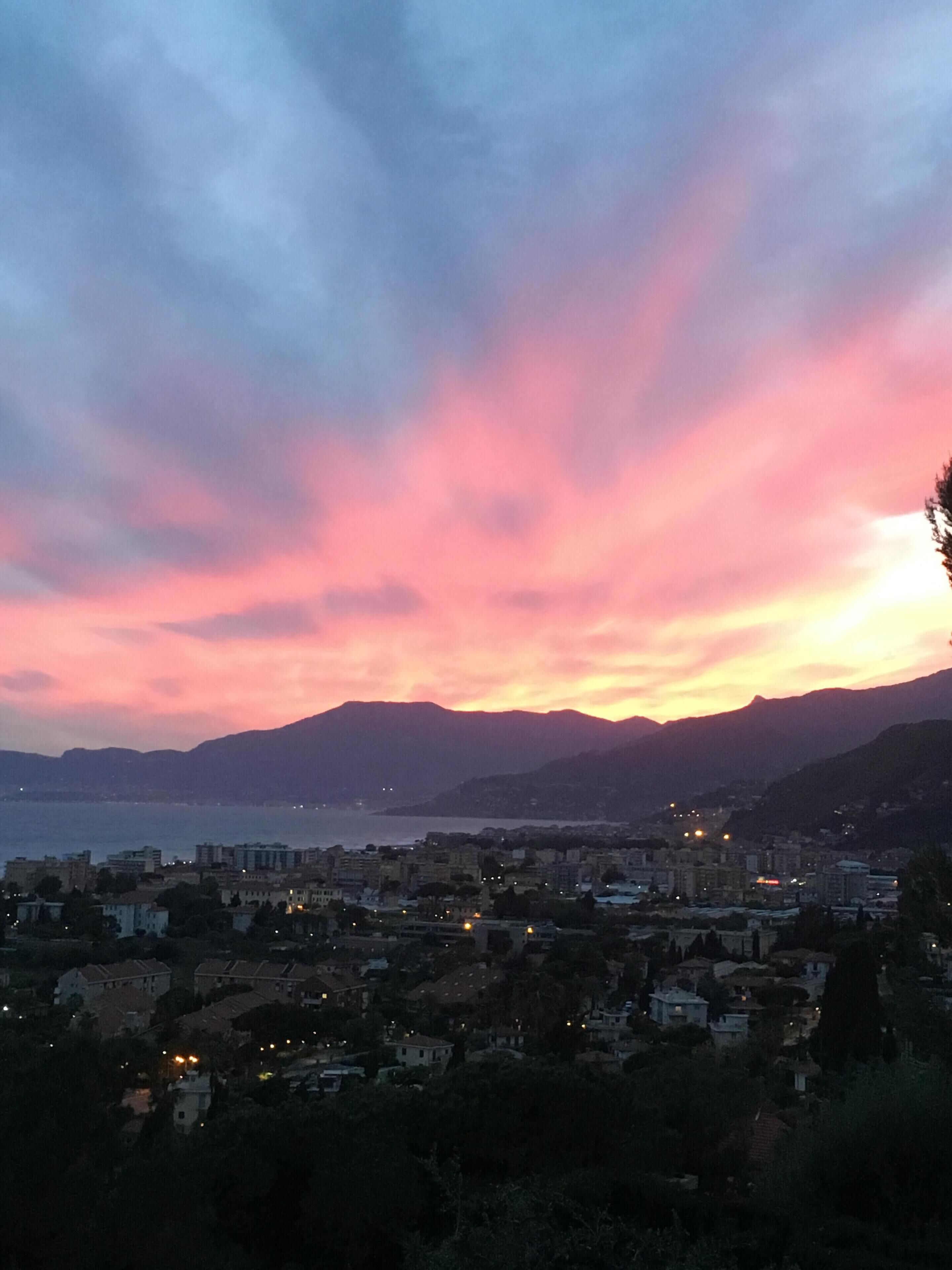 Paesaggio costiero al crepuscolo con cielo rosa e viola, montagne sullo sfondo e edifici lungo la riva.