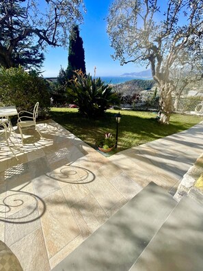 Un patio con mobiliario exterior, un pequeño jardín y vistas al mar y colinas lejanas.
