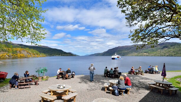 A lakeside picnic area with wooden tables and benches, a boat on the water, and hills in the background. - The Bridge Suite, Invermoriston - The Great Glenway (Invermoriston)