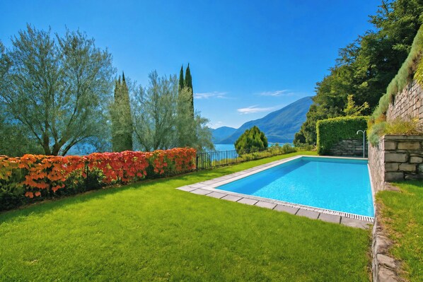 A rectangular swimming pool with a stone border, surrounded by well-maintained grass and a hedge with red flowers. - Villa San Michele, Griante - Cadenabbia, Italy (Griante - Cadenabbia)