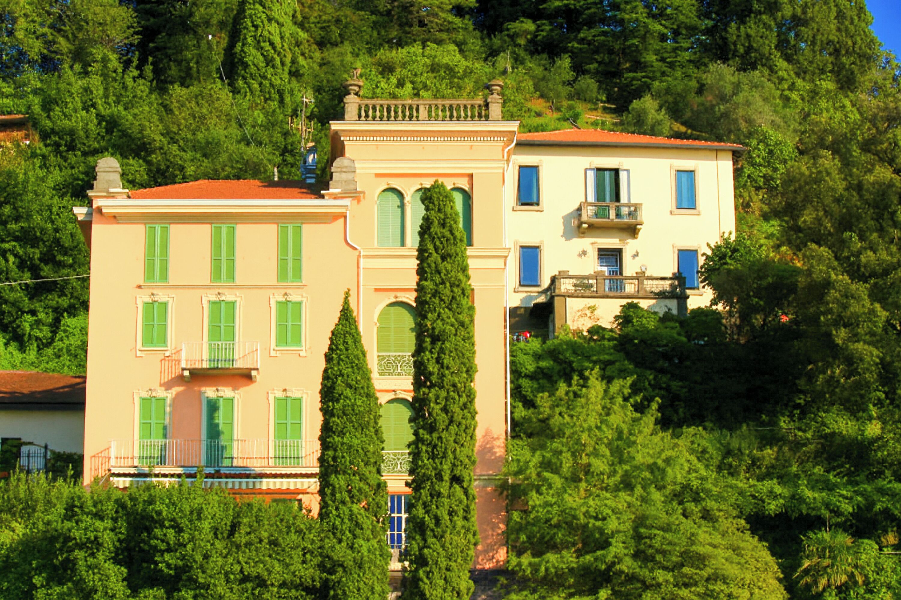 A multi-story building with a red roof, green shutters, and a balcony. It is surrounded by lush greenery and trees.