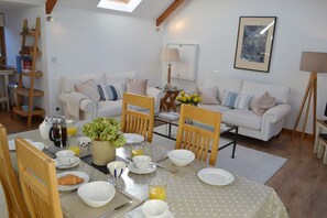 Ein Essbereich mit einem für das Frühstück gedeckten Tisch, einschließlich Tellern, Tassen und einem Croissant. Zwei Holzstühle, eine weiße Couch und ein Couchtisch mit Blumen stehen dort.