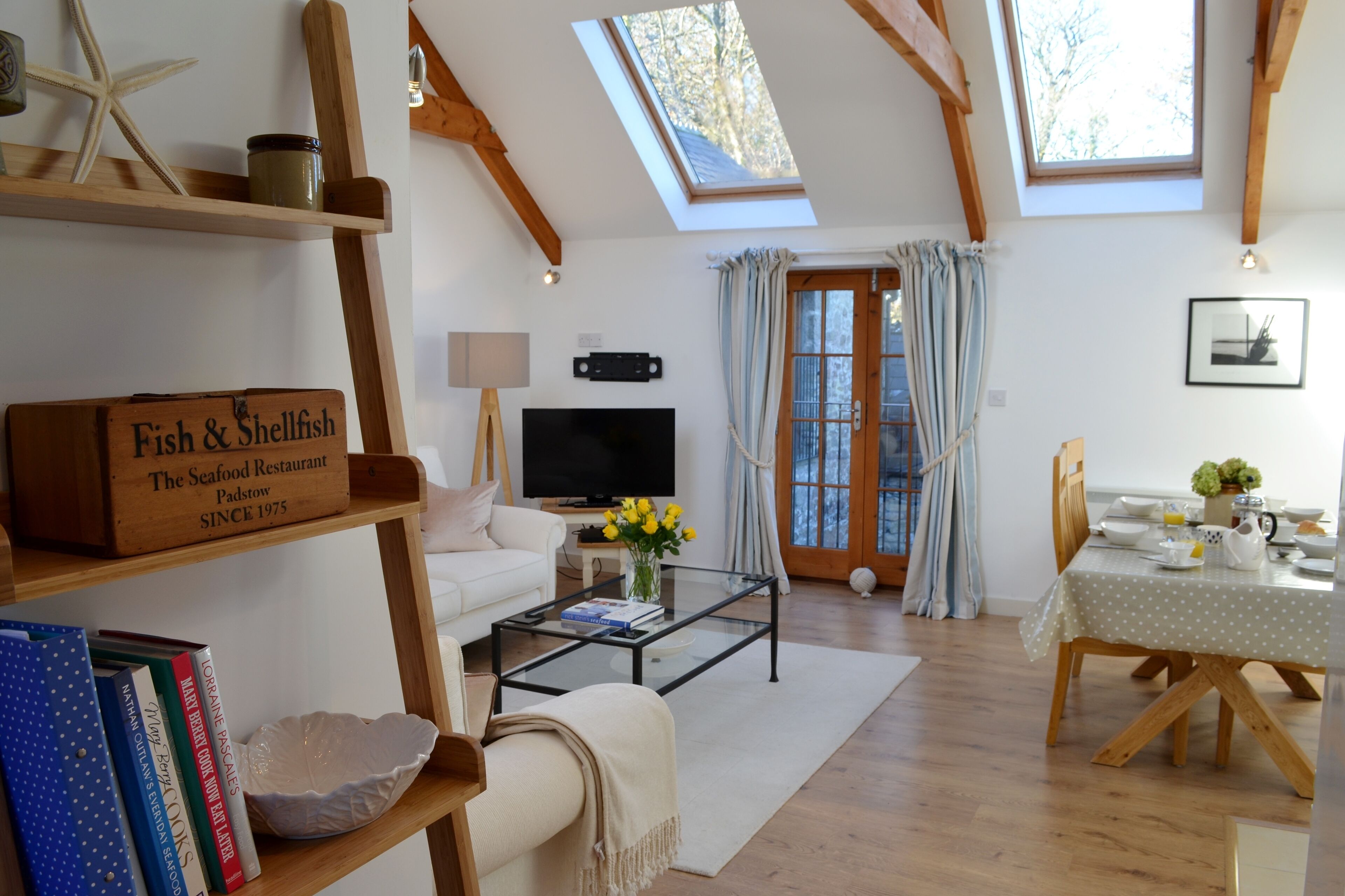 Una sala de estar acogedora con televisor, libros y una mesa de centro. Hay una zona de comedor con una mesa dispuesta para dos personas.