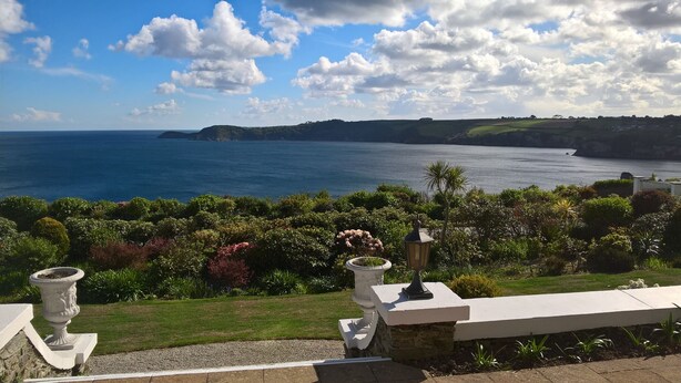 A coastal landscape with a view of the sea, lush greenery, and a stone pathway leading to a seating area.