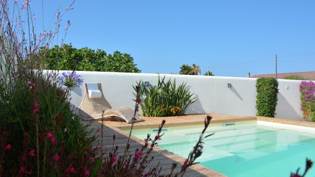 A swimming pool with a wooden deck, surrounded by lush greenery and flowering plants. - Modern Villa with pool, sea view and mediterranean garden (Bari)