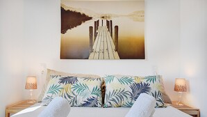 Une chambre d’hôtel avec un lit, deux oreillers ornés d’un motif de feuilles tropicales, deux lampes de chevet et un tableau représentant une jetée menant à une montagne.