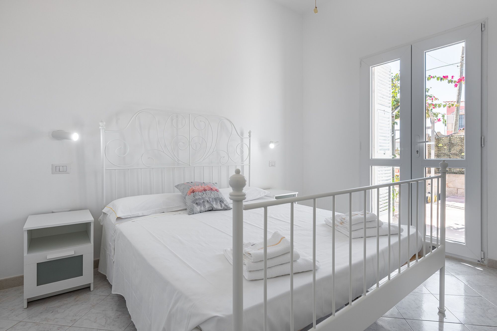 A white bedroom with a bed, nightstand, and a balcony with a view of the outside.