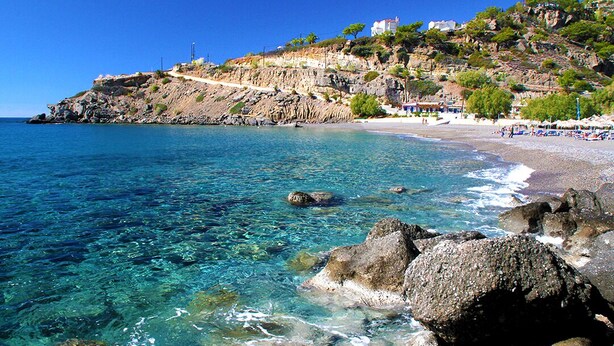 Una playa costera con aguas cristalinas, costa rocosa y edificios en una colina.
