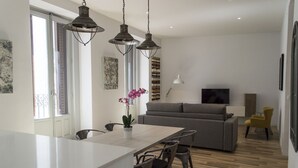 A modern kitchen with a central island, dining chairs, pendant lights, a sofa, a TV, and artwork on the walls.