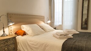 A neatly made bed with white linens, a grey throw blanket, and folded towels on the side. There is a bedside lamp on the left and a nightstand with a lamp and a decorative item.