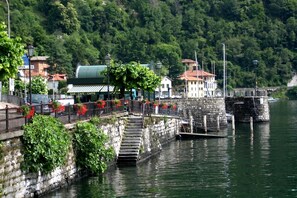 A waterfront area with a stone staircase leading down to a dock, surrounded by greenery and buildings with red flowers.