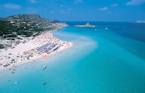 A coastal beach with clear turquoise waters, a sandy shore, and a mountainous backdrop.
