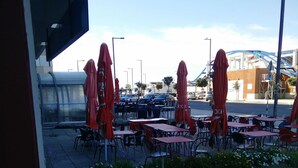 Ein Straßencafé mit Tischen und Stühlen, das mit roten Sonnenschirmen beschattet wird, mit Blick auf die Straße und die Gebäude im Hintergrund.