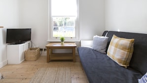 Et lille, lyst værelse med fjernsyn, sofa, vindue og et lille bord med en plante.
