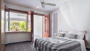 A bedroom with a bed, a ceiling fan, a window with a view of greenery, and a patterned floor.