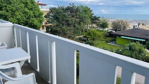 Un balcon avec une balustrade blanche, une petite table et deux chaises blanches. En arrière-plan, on aperçoit une plage et un bâtiment vert.