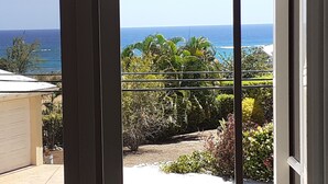 A view through a window of a garage and a garden with tropical plants and a clear blue sky. - Ocean view apartment with plunge pools, roof terrace, balcony and near the beach (Enterprise)