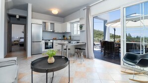 Eine moderne Küche mit weißen Schränken, ein Essbereich mit rundem Tisch und Blick auf einen Hügel durch große Glasschiebetüren.
