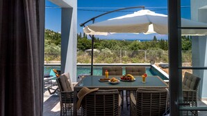 Eine Terrasse im Freien mit Korbmöbeln, einem Tisch mit Frühstücksangeboten und Blick auf einen Swimmingpool und Grünflächen.