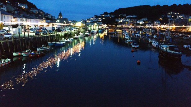 Un port au crépuscule, avec des bateaux amarrés, des bâtiments sur les collines et un beffroi.