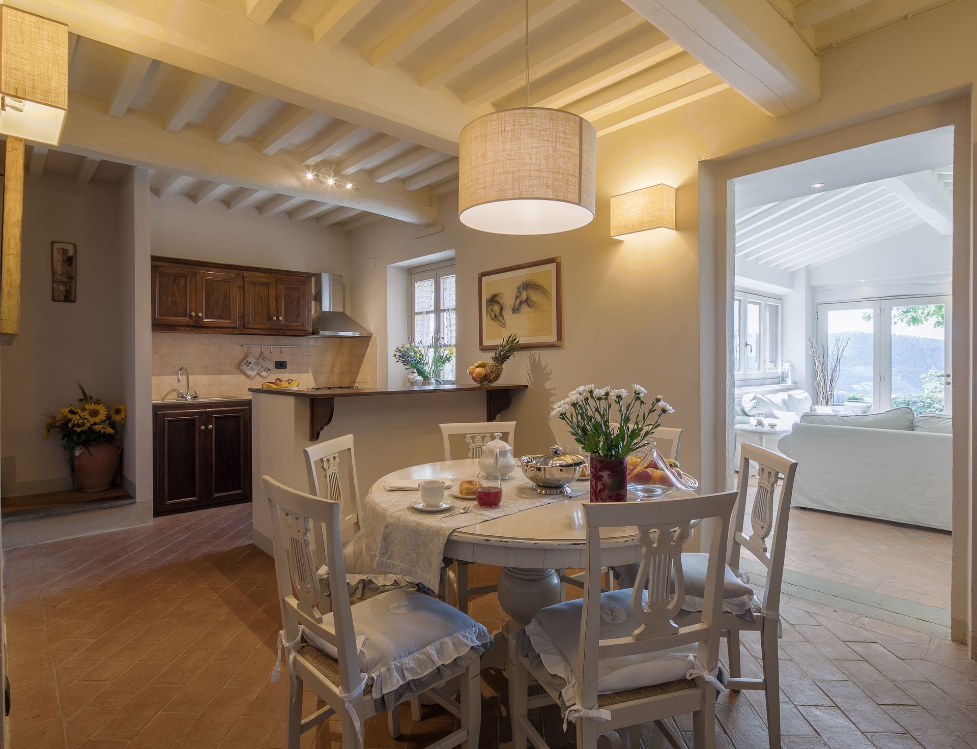 A dining area with a round table, white chairs, and a vase of flowers. There is a kitchen in the background with wooden cabinets and a window.