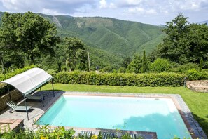 Una piscina rectangular con borde de piedra, rodeada por un césped y un seto.